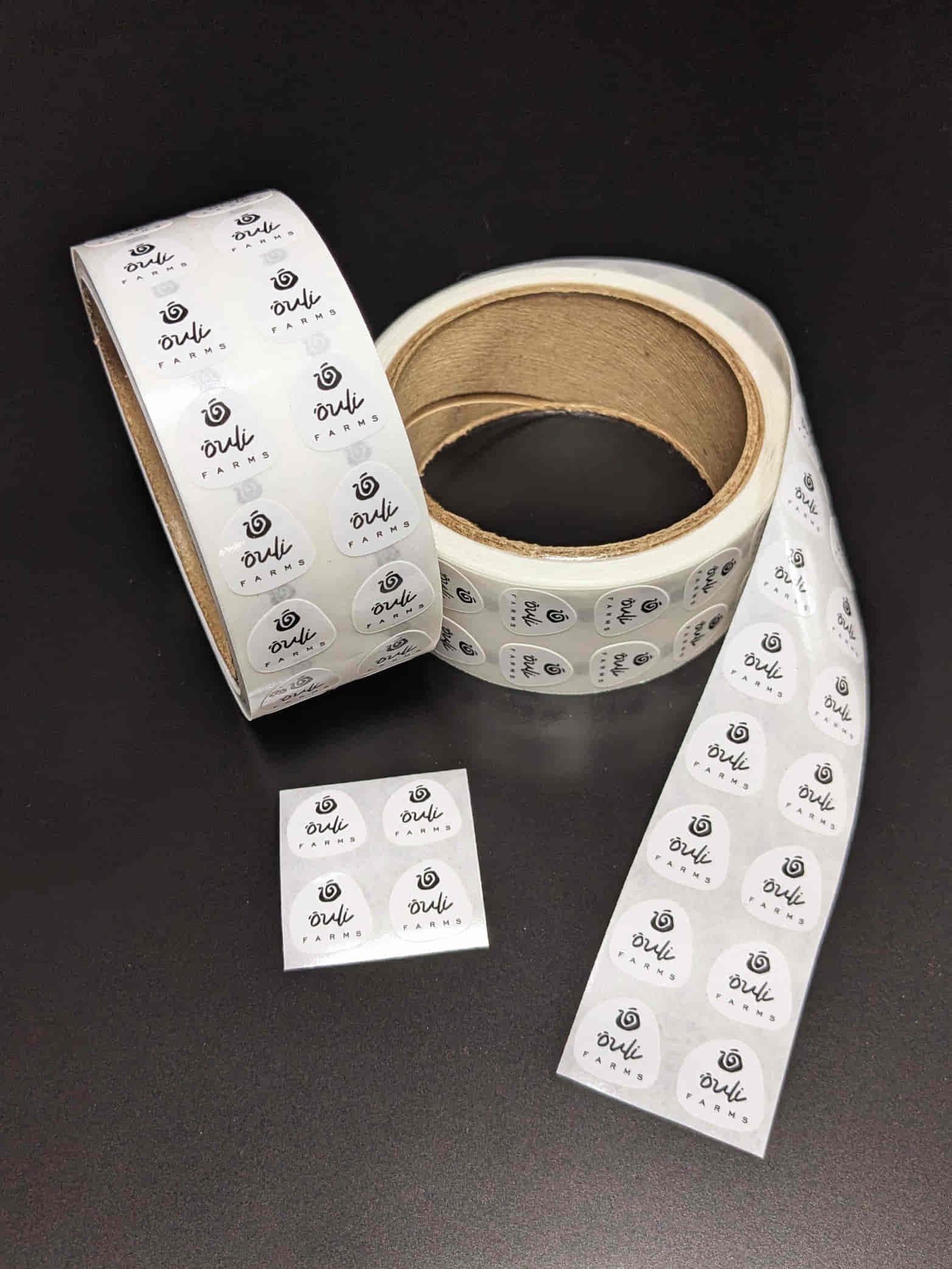 Two rolls of circular "Orli Farms" labels with a glossy finish, partially unrolled, against a dark background.