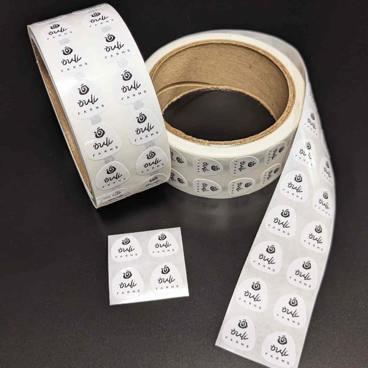 Two rolls of circular "Orli Farms" labels with a glossy finish, partially unrolled, against a dark background.